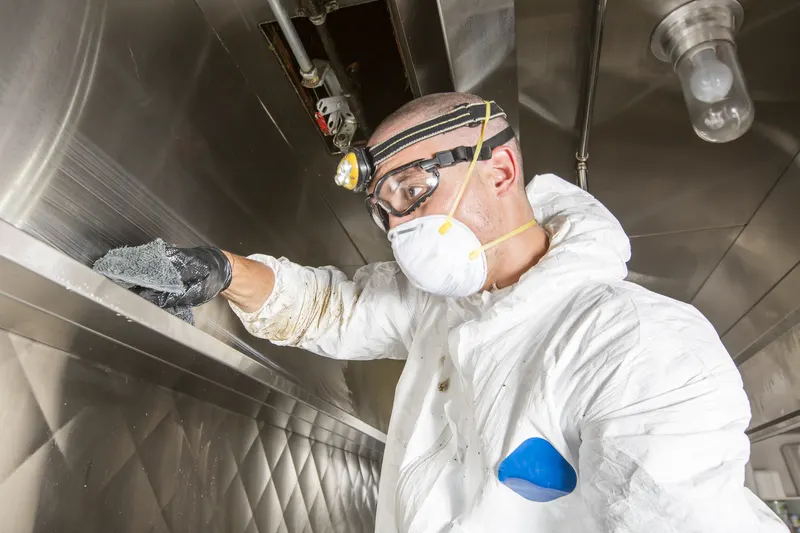 Technician in goggles and headlamp wiping the front edge of a stainless exhaust hood with a grey cloth
