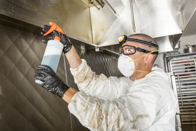 Technician in respirator and goggles raising a spray bottle to the underside of a stainless exhaust canopy