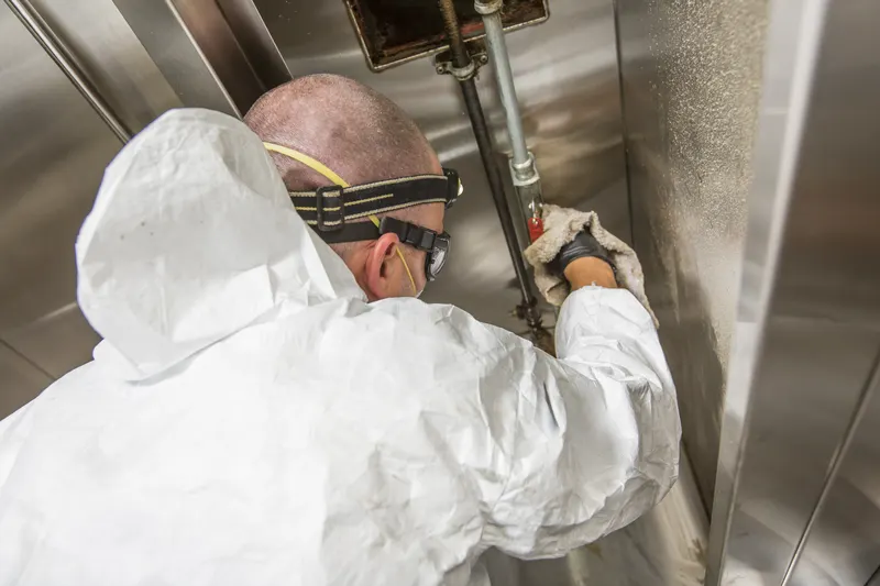 Technician in a hooded coverall wiping the plenum wall beside the suppression pipework inside an exhaust hood