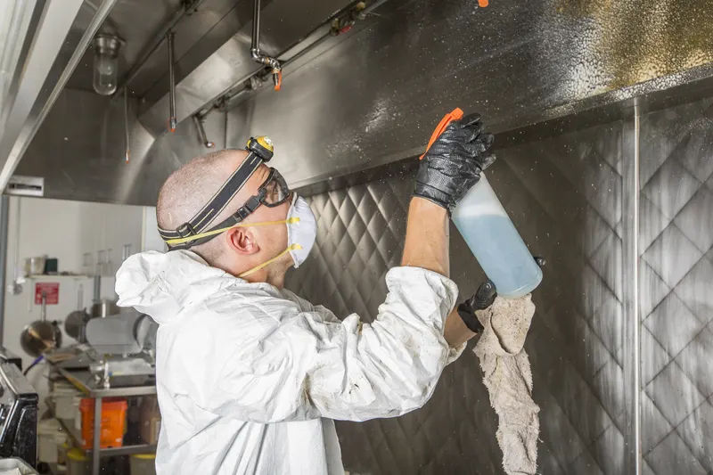 Technician spraying and wiping the inside of a stainless exhaust hood with commercial cooking equipment on the line behind